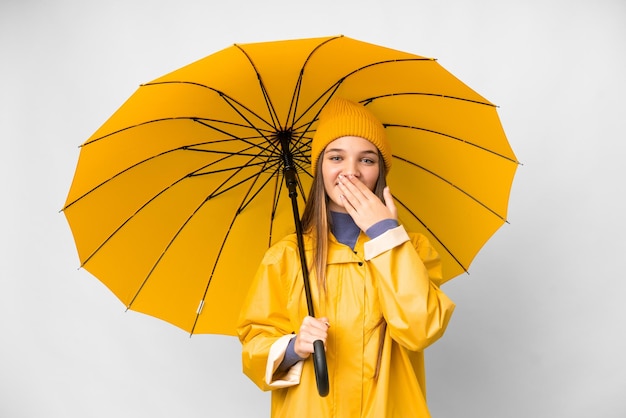 Teenager girl with rainproof coat and umbrella over isolated white background happy and smiling covering mouth with hand