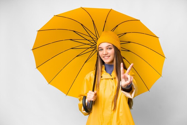 防雨コートと分離の白い背景の上の傘を持つティーンエイ ジャーの女の子笑顔と勝利のサインを示す