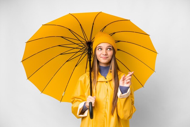 Девушка-подросток в непромокаемом пальто и зонтике на изолированном белом фоне, скрещивая пальцы и желая всего наилучшего