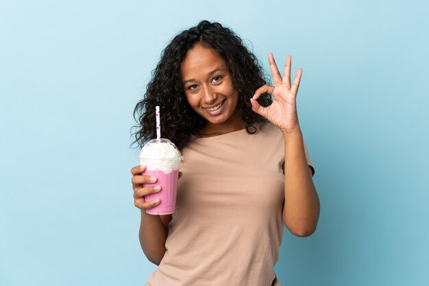 Девушка-подросток с клубничным молочным коктейлем изолирована на синем фоне, показывая пальцами знак ОК