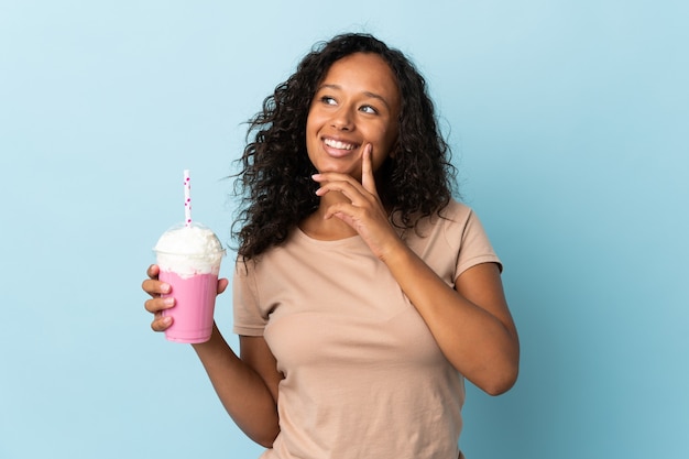 Teenager girl  with strawberry milkshake isolated on blue background thinking an idea while looking up