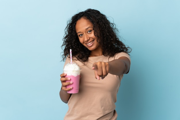 Девушка-подросток с клубничным молочным коктейлем изолирована на синей стене, указывая вперед с счастливым выражением лица