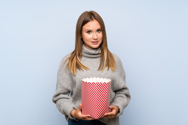 Teenager girl with sweater over isolated blue background holding a bowl of popcorns