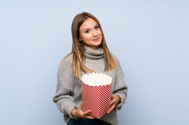 Teenager girl with sweater over isolated blue background holding a bowl of popcorns