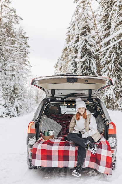 クリスマスロードトリップアドベンチャーと地元旅行のコンセプトのために装飾された車のトランクに座っている魔法瓶を持つティーンエイジャーの女の子雪に覆われた森での冬のピクニック新年の休暇