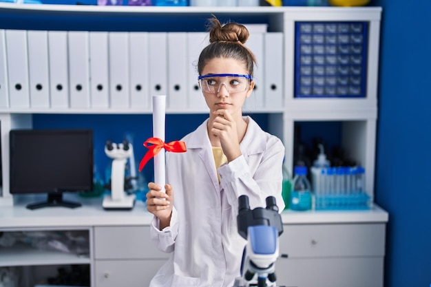Teenager girl working at scientist laboratory holding degree serious face thinking about question with hand on chin thoughtful about confusing idea
