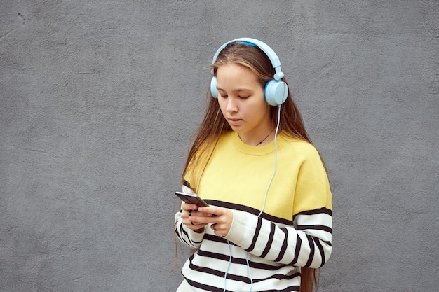 Teenager girl in a yellow striped sweater uses a smartphone and headphones listens to music on a gray background