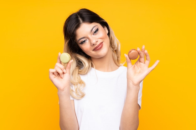 Teenager girl on yellow wall offering colorful French macarons