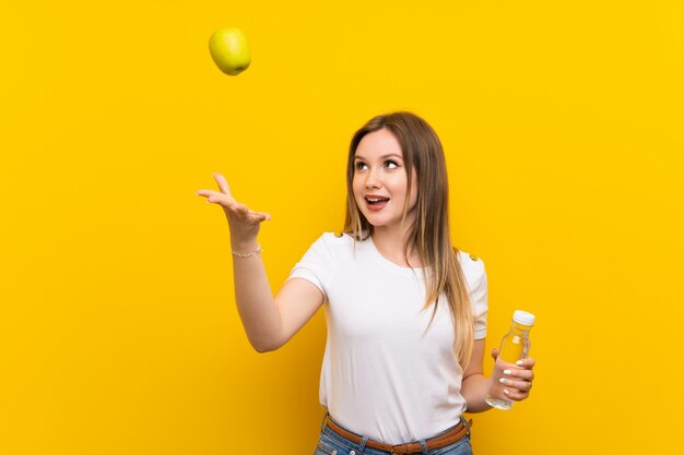 Teenager girl over yellow wall with an apple and a bottle of water