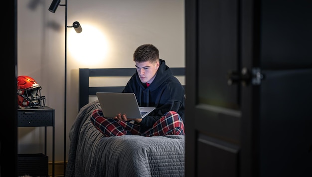 A teenager guy sits in a room on a bed and uses a laptop