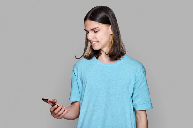 Teenager guy with smartphone in his hands on gray colored background Smiling happy teenage male looking at phone screen