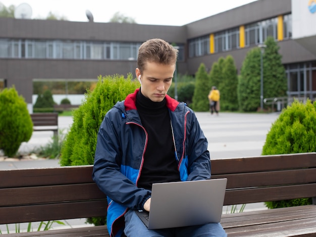 ヘッドホンをつけた10代の学生が、大学近くの公園でノートパソコンを持ってベンチに座っています。