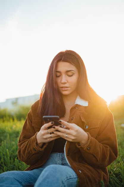 A teenager holding her phone while sitting at a green meadow during the sunset in spring