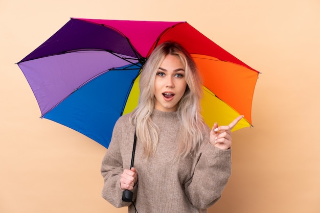 Teenager holding an umbrella surprised and pointing finger to the side