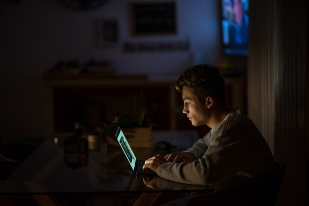 Teenager at home using his laptop to do homework in video call with his class or playing videogames at night - young man smiling surfing on the net in quarantine  in his house