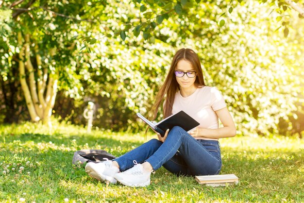 teenager is sitting on the grass studying a student girl under a tree in the university park with a backpack and textbooks