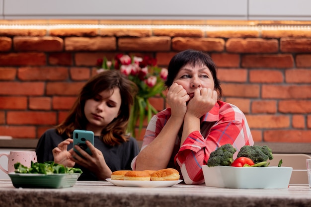 Teenager is sitting with the phone next to hir grandmother and does not pay attention to her