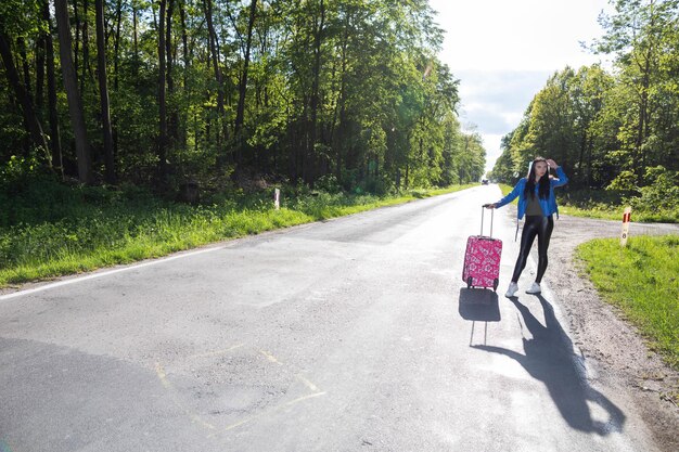 A teenager is standing with a pink suitcase on wheels and tries to stop the opportunity to go on vacation faster