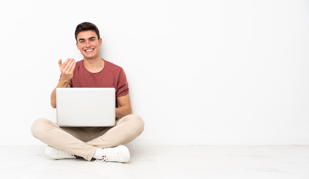 Teenager man sitting on the flor with his laptop inviting to come with hand. Happy that you came