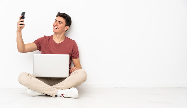 Uomo dell'adolescente che si siede su flor con il suo computer portatile che fa un selfie