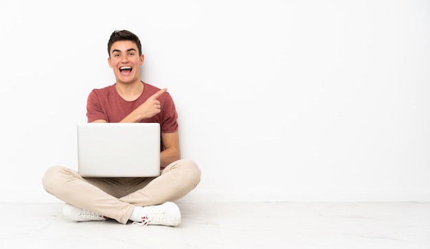 Teenager man sitting on the flor with his laptop surprised and pointing side