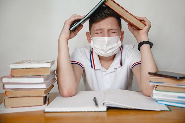 A teenager in a mask at home sitting at lessons covers his head with textbooks. Coronavirus concept