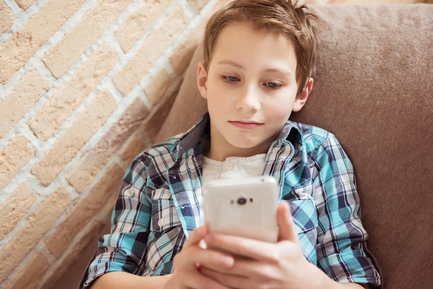 Teenager messaging to friends chat on mobile lying on couch in living room at home