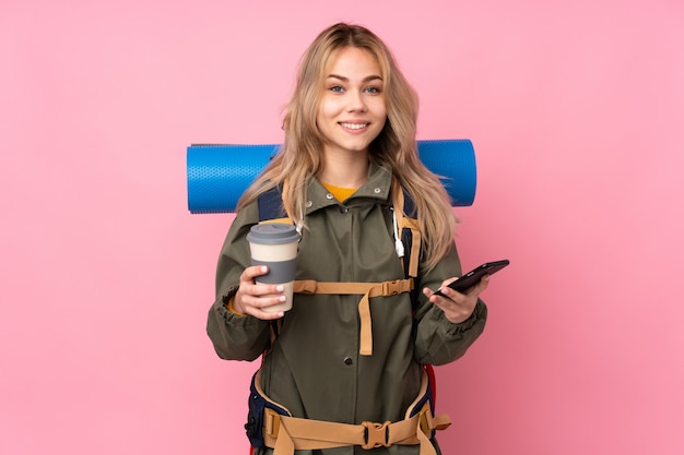 Teenager mountaineer girl with a big backpack isolated on pink wall holding coffee to take away and a mobile