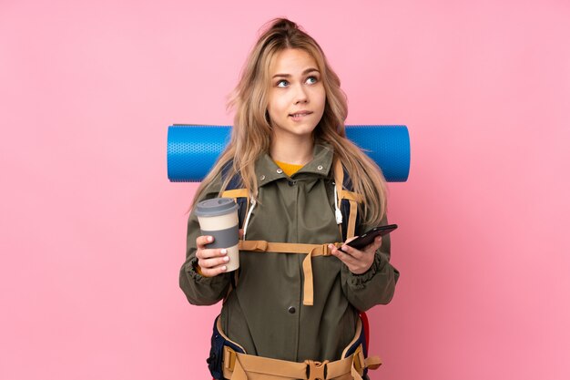 Teenager mountaineer girl with a big backpack on pink holding coffee to take away and a mobile while thinking something