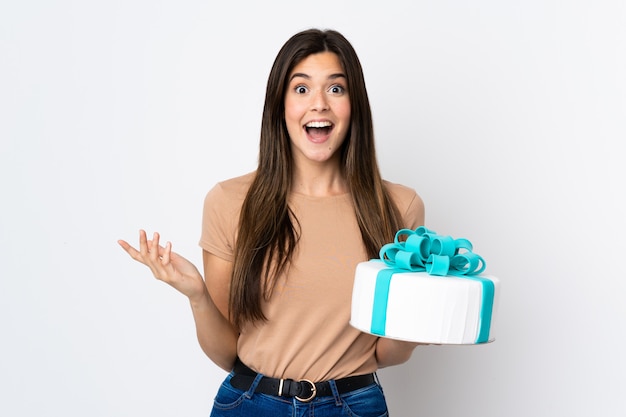 Teenager pastry chef holding a big cake over isolated white background with shocked facial expression