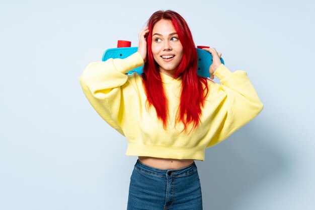 Teenager red hair girl isolated on blue wall with a skate and looking up