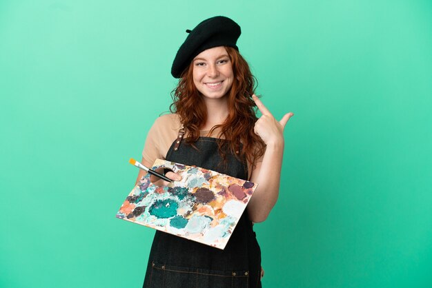 Teenager redhead artist holding a palette isolated on green background giving a thumbs up gesture