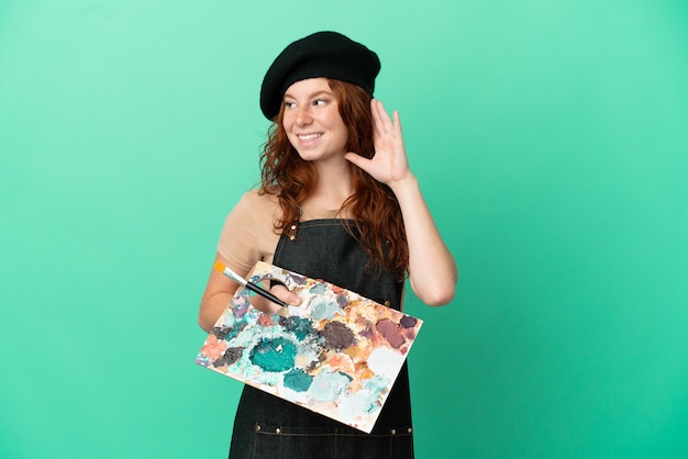 Teenager redhead artist holding a palette isolated on green background listening to something by putting hand on the ear