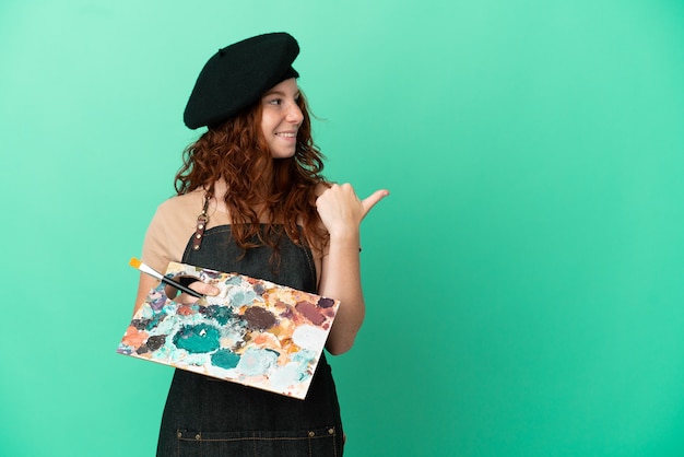 Foto artista adolescente dai capelli rossi che tiene una tavolozza isolata su sfondo verde che punta al lato per presentare un prodotto