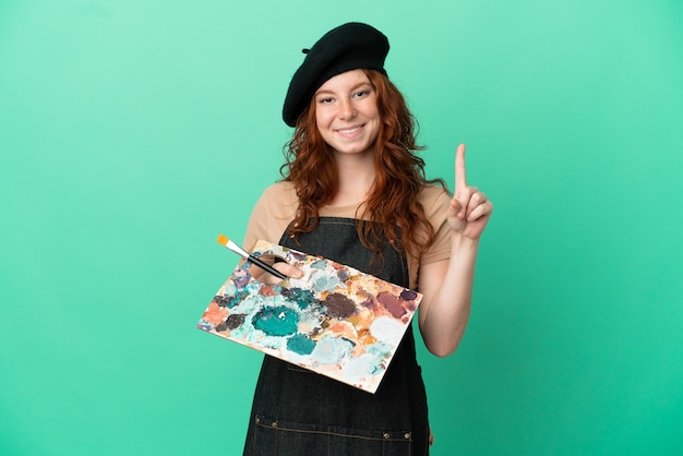Foto artista adolescente dai capelli rossi che tiene una tavolozza isolata su sfondo verde che mostra e solleva un dito in segno del meglio