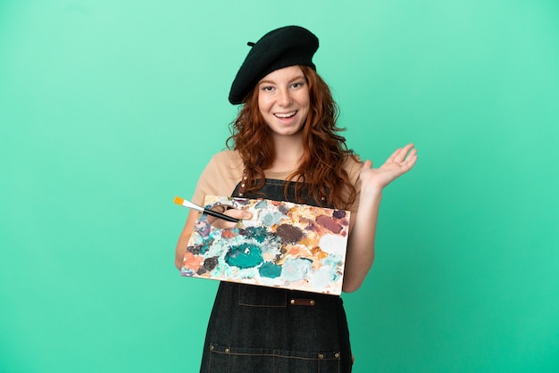 Teenager redhead artist holding a palette isolated on green background with shocked facial expression