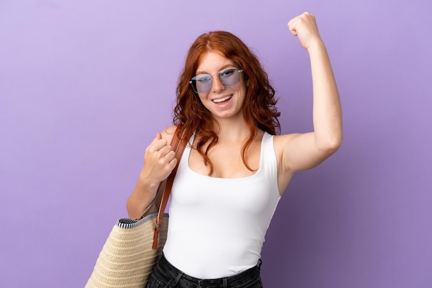 Teenager redhead girl holding a beach bag isolated on purple background celebrating a victory