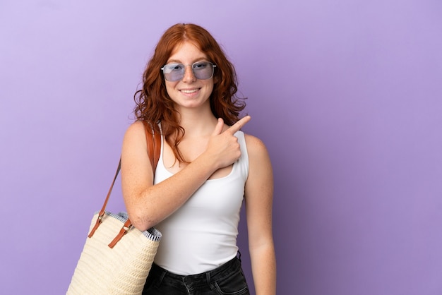 Teenager redhead girl holding a beach bag isolated on purple background pointing to the side to present a product