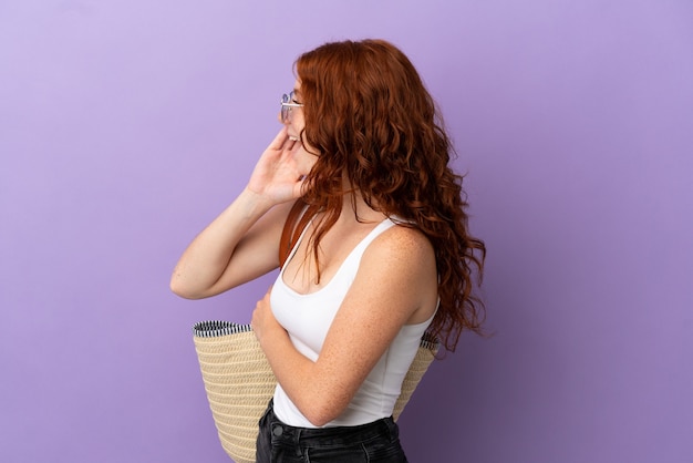 Teenager redhead girl holding a beach bag isolated on purple background shouting with mouth wide open to the lateral