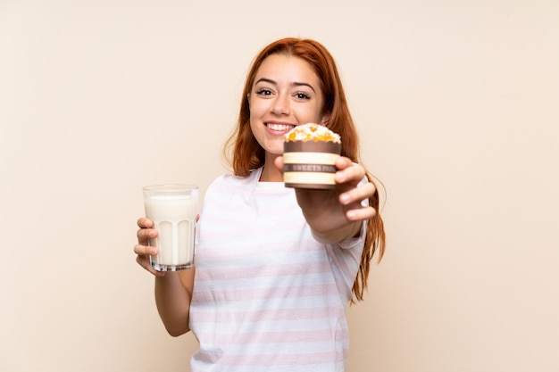 Teenager redhead girl holding a glass of milk and a muffin over isolated background