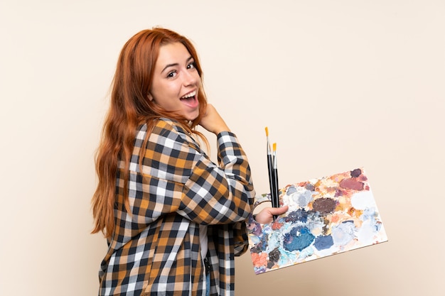 Teenager redhead girl holding a palette over isolated wall celebrating a victory