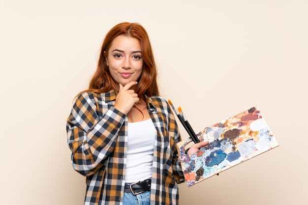 Teenager redhead girl holding a palette over isolated wall laughing