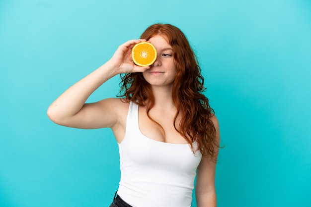 Teenager redhead girl isolated on blue background in swimsuit and holding an orange