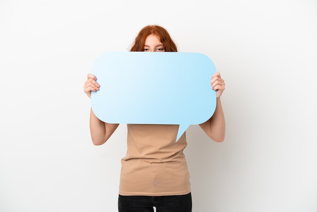 Photo teenager redhead girl isolated on white background holding an empty speech bubble hiding behind it hiding behind it