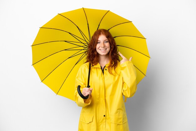 10代の赤毛の女の子の防雨コートと傘は、親指を立てるジェスチャーを与える白い背景で隔離