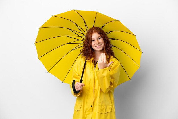 Teenager redhead girl rainproof coat and umbrella isolated on white background inviting to come with hand. Happy that you came