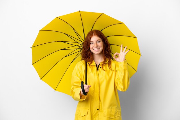 10代の赤毛の少女の防雨コートと傘は白い背景で隔離指でOKサインを示しています