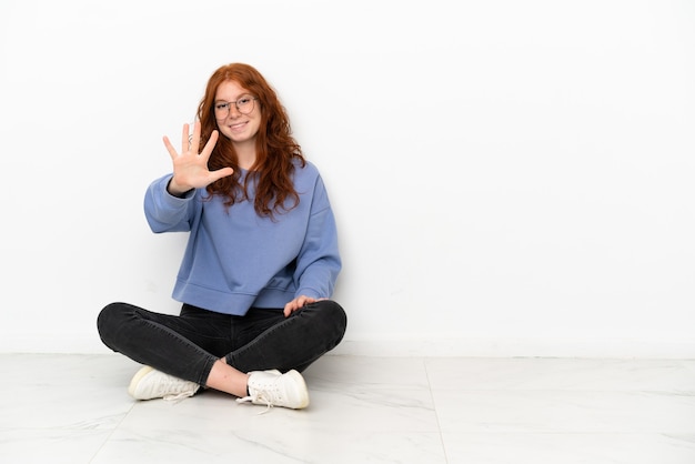 Ragazza rossa dell'adolescente che si siede sul pavimento isolata su fondo bianco che conta cinque con le dita