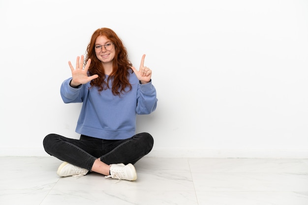 Рыжая девушка-подросток сидит на полу на белом фоне, считая семь пальцами