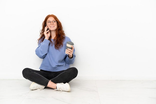 흰색 배경에 격리된 바닥에 앉아 있는 10대 빨간 머리 소녀는 테이크아웃 커피와 모바일을 들고 있습니다.